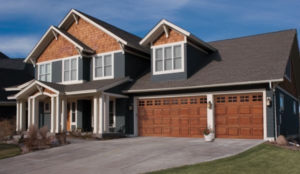 Two-story blue house with brown wood accents, two car garage, and concrete driveway on a sunny day.