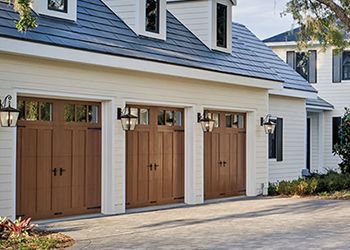 Three brown garage doors on a white house with black lanterns and a paved driveway.