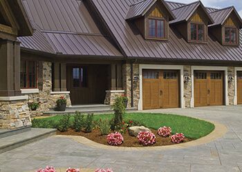 House exterior with brown metal roof, stone pillars, and wooden garage doors. Landscaping with pink flowers.