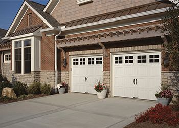 Two white garage doors with square windows, beneath a pergola, in front of a brick and stone house.