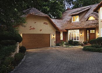 House with thatched roof, brown garage door, cobblestone driveway, and lush landscaping.