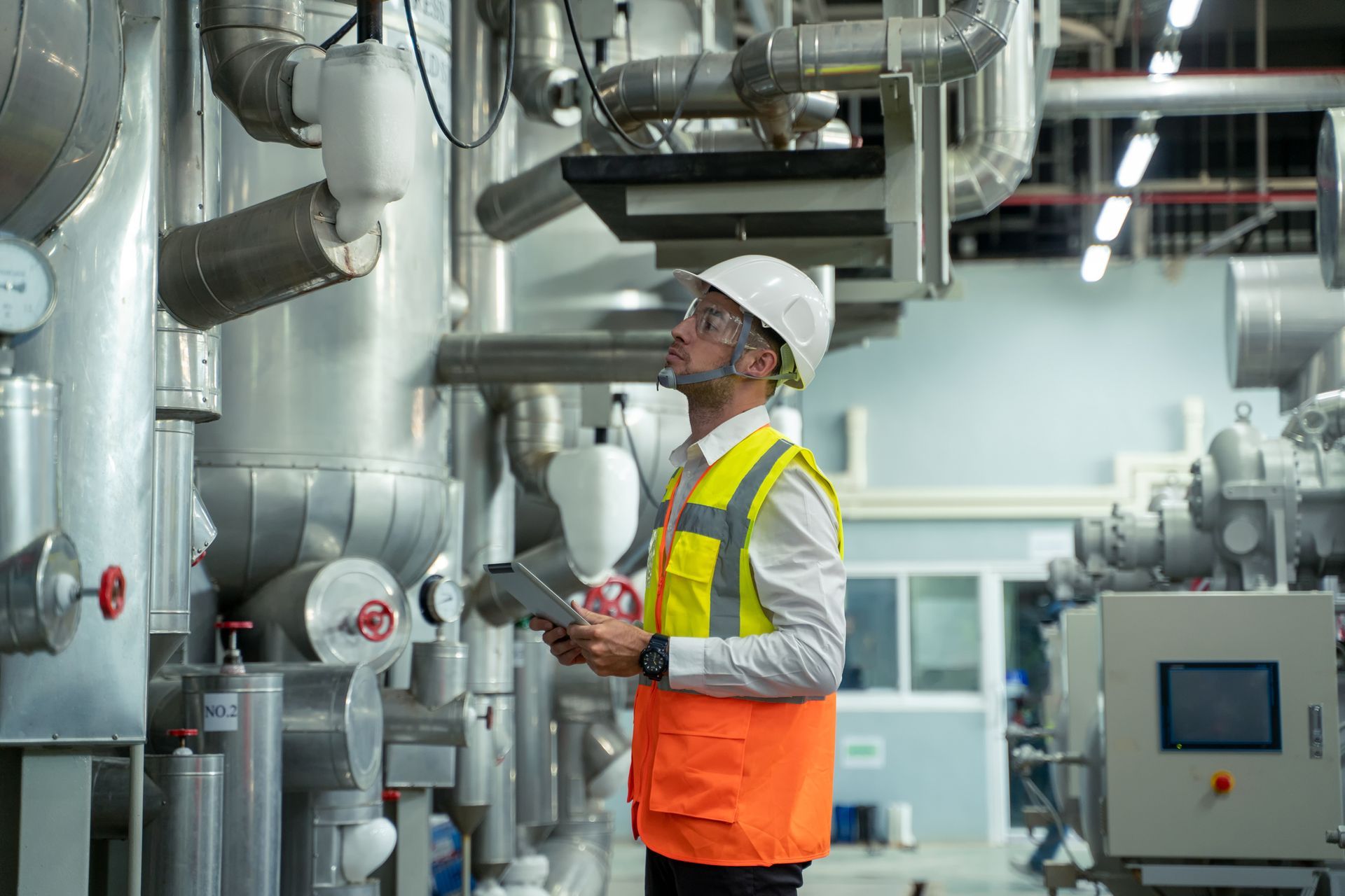 Engineer in safety vest and hard hat inspects pipes in a factory, holding a tablet.