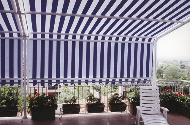 Tenda a righe blu e bianche sopra un patio con piante in vaso e sedie bianche impilate, con vista su un panorama nebbioso.