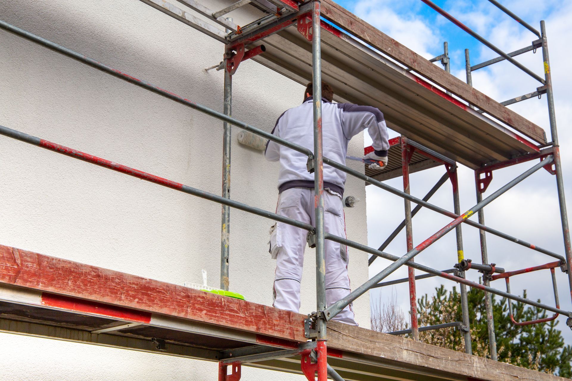 Ein Mann steht in Arbeitskleidung auf einem Gerüst und streicht mit einer Malerrolle die Fassade.