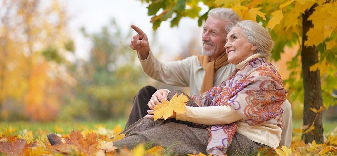 An elderly couple is sitting in the leaves in a park.