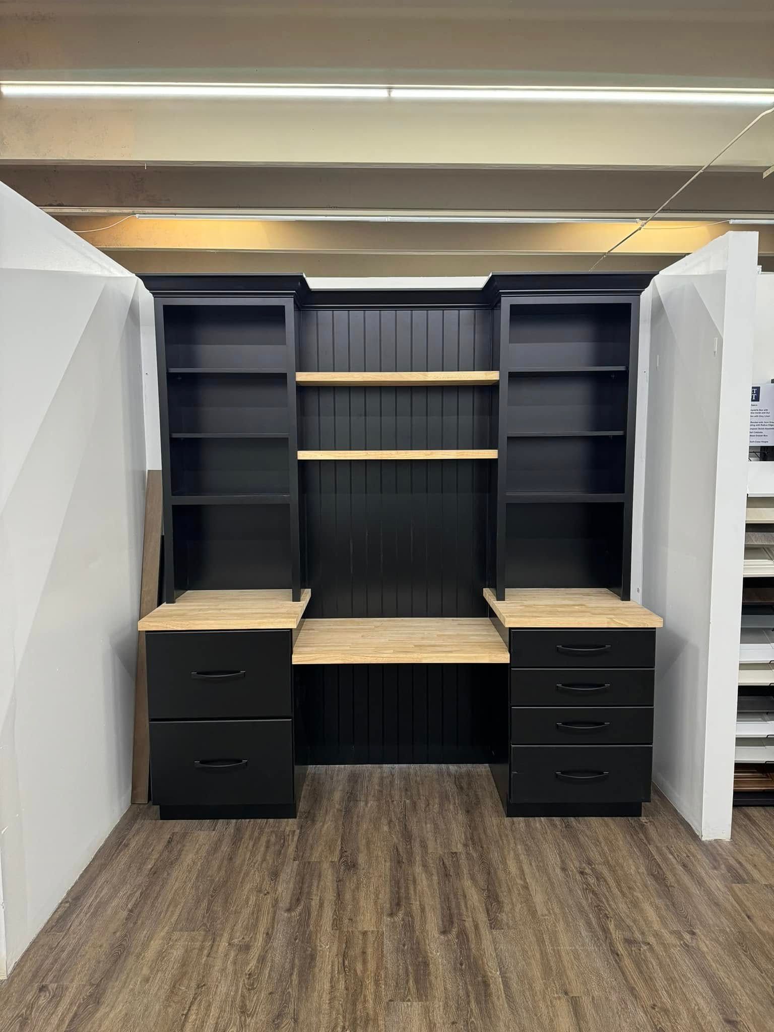 Black built-in desk with bookshelves, light wood countertop and backing, on a brown floor.