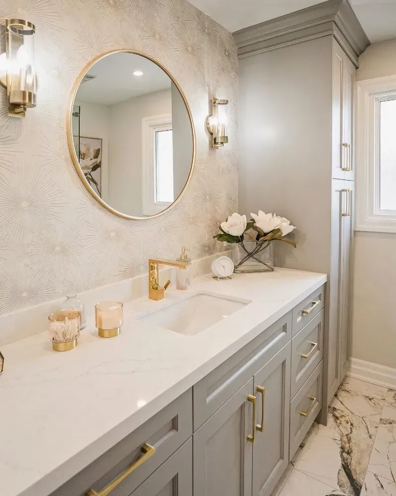 Bathroom vanity with a round mirror, gold accents, gray cabinets, and a white countertop.