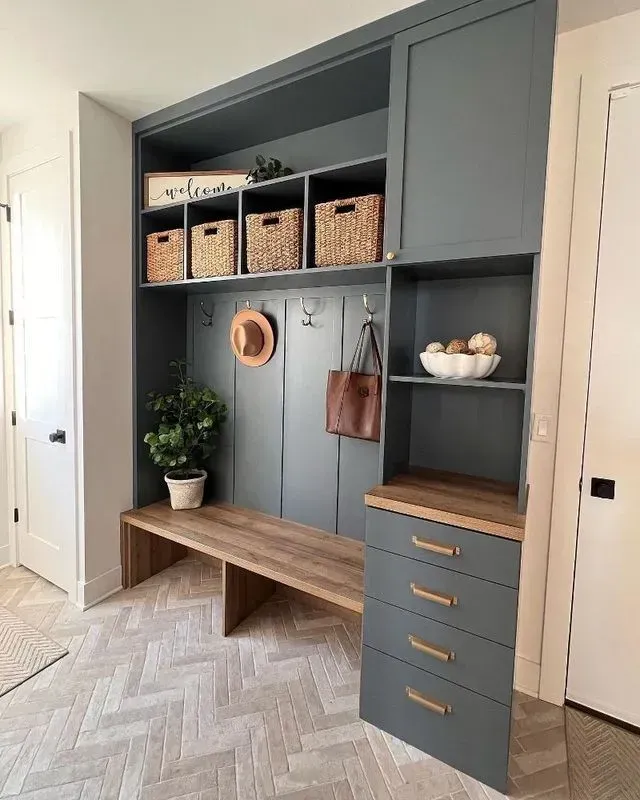 Built-in mudroom with bench, storage, and hooks, painted blue-gray. Wicker baskets, a hat, and a purse hang inside.