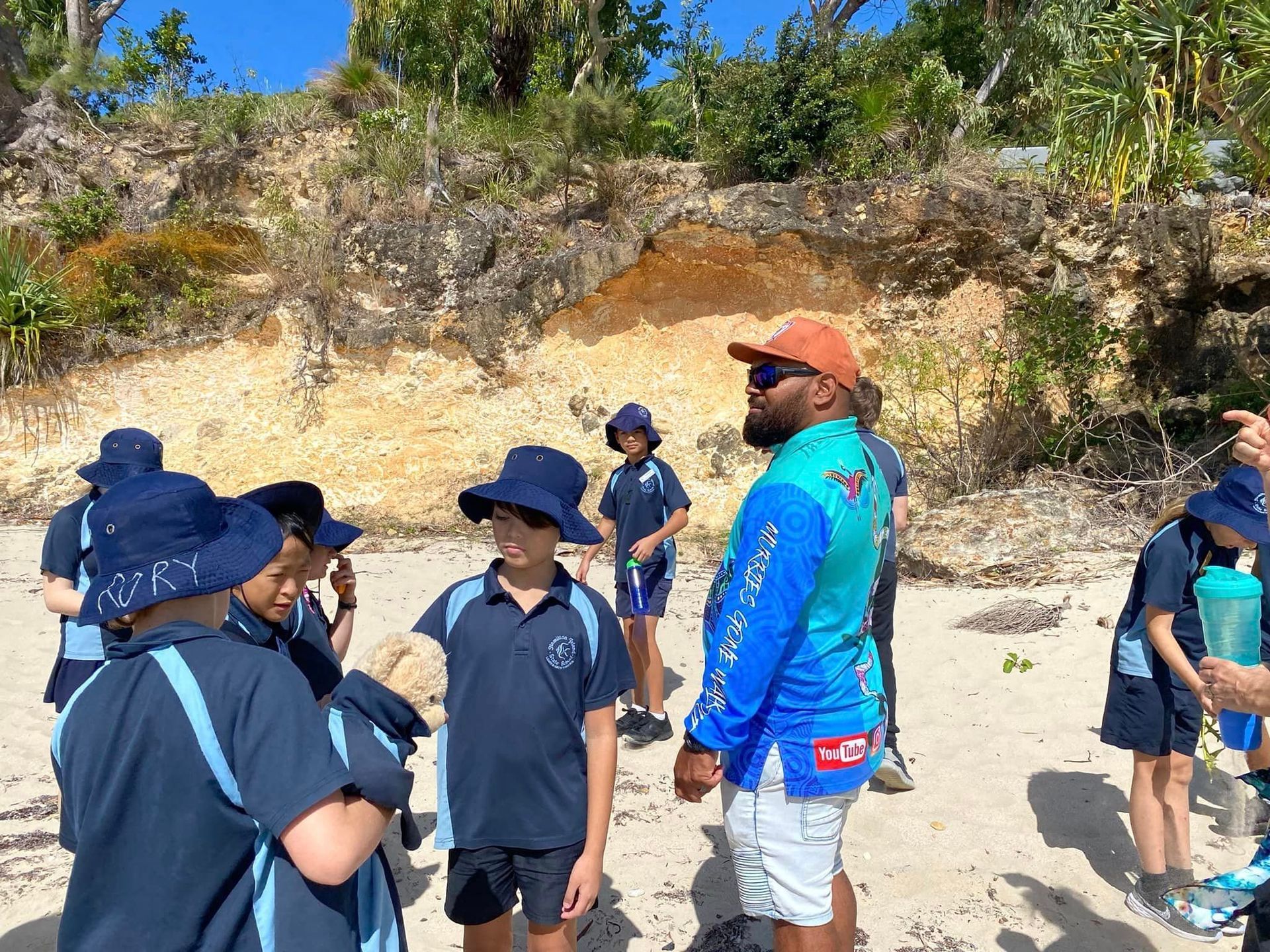A group of children are standing on a beach talking to a man.
