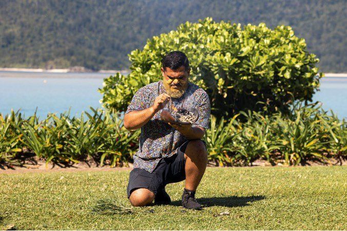 A man with a beard is kneeling on the grass in front of a body of water.