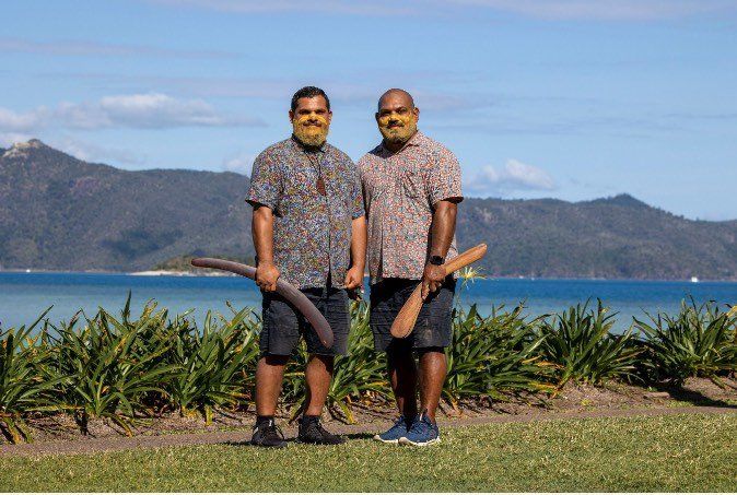 Two men are standing next to each other holding boomerangs in front of a body of water.