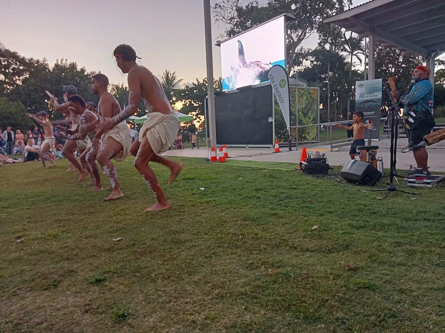 A group of men are dancing in a field in front of a large screen.