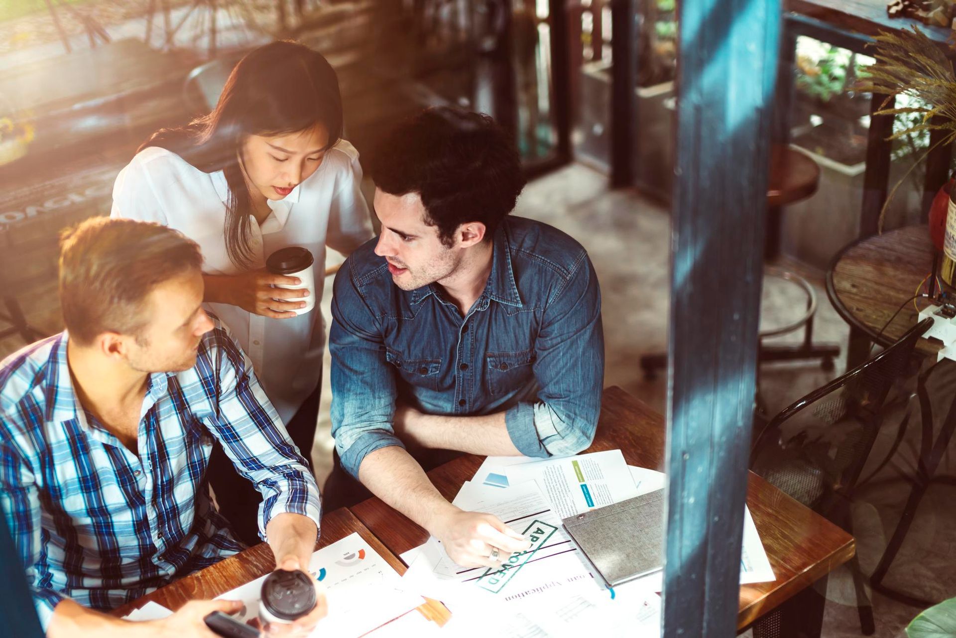 Three coworkers reviewing papers together at a cafe table, with a laptop and coffee cups.