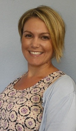Smiling woman with blonde hair in a patterned top and light blue cardigan against a plain gray wall
