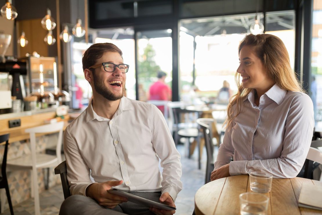 Two people chatting and smiling at a café table with drinks and newspapers.