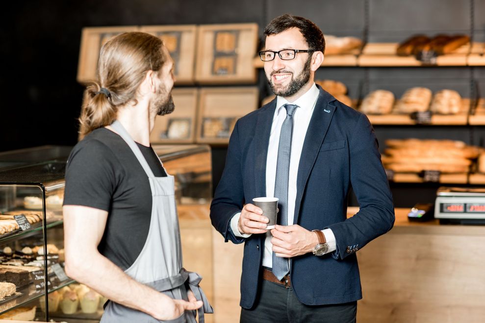 Two coworkers talking in a bakery, one holding a coffee cup, shelves of bread in the background