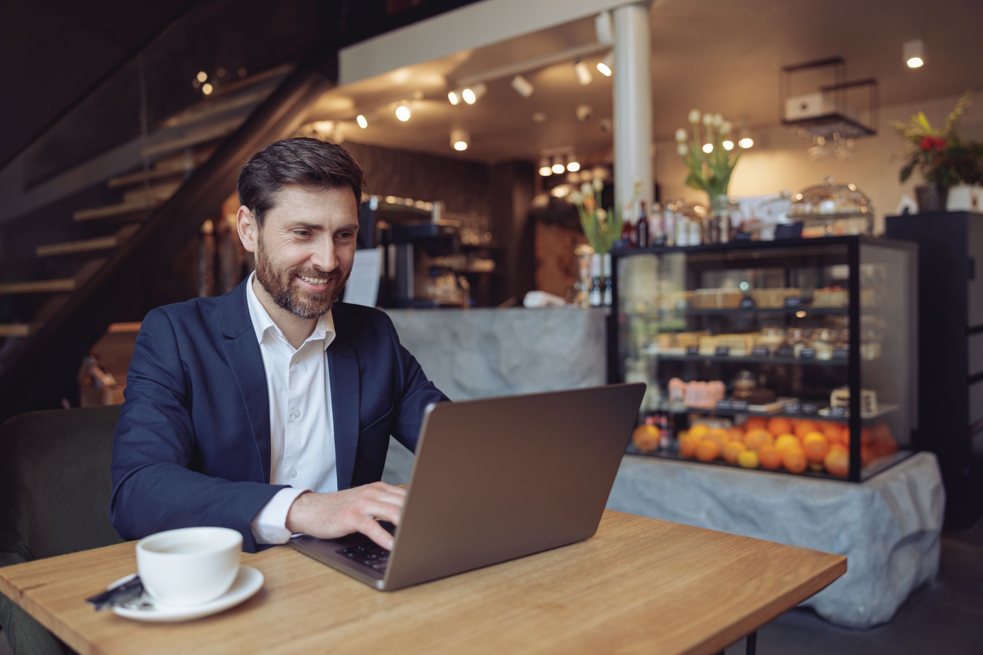 Man in a navy blazer using a laptop at a café table with a coffee cup nearby