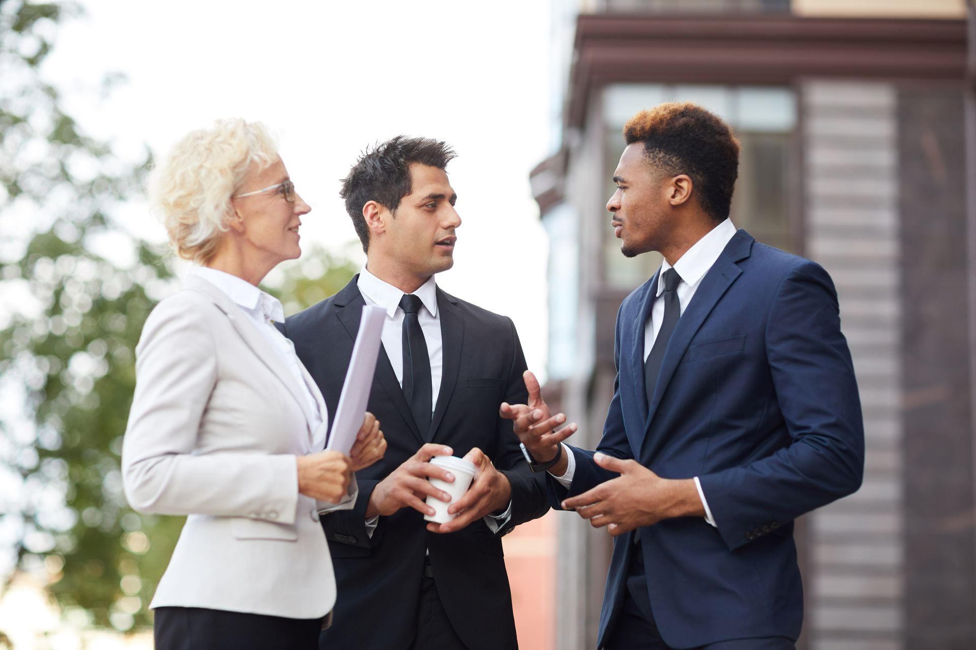 Three businesspeople in suits talking outdoors near an office building