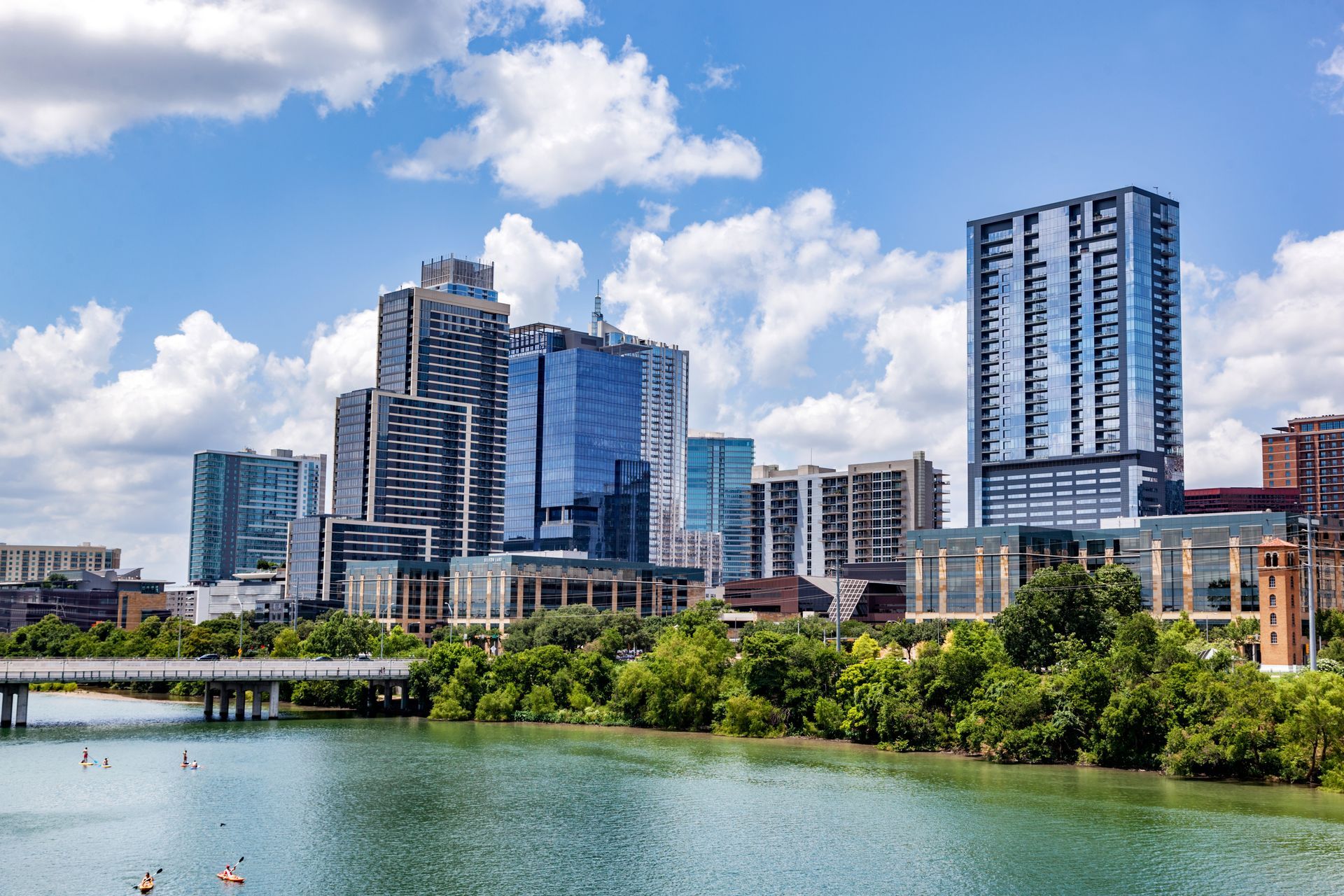 City skyline with modern high-rises beside a calm river under a bright blue sky