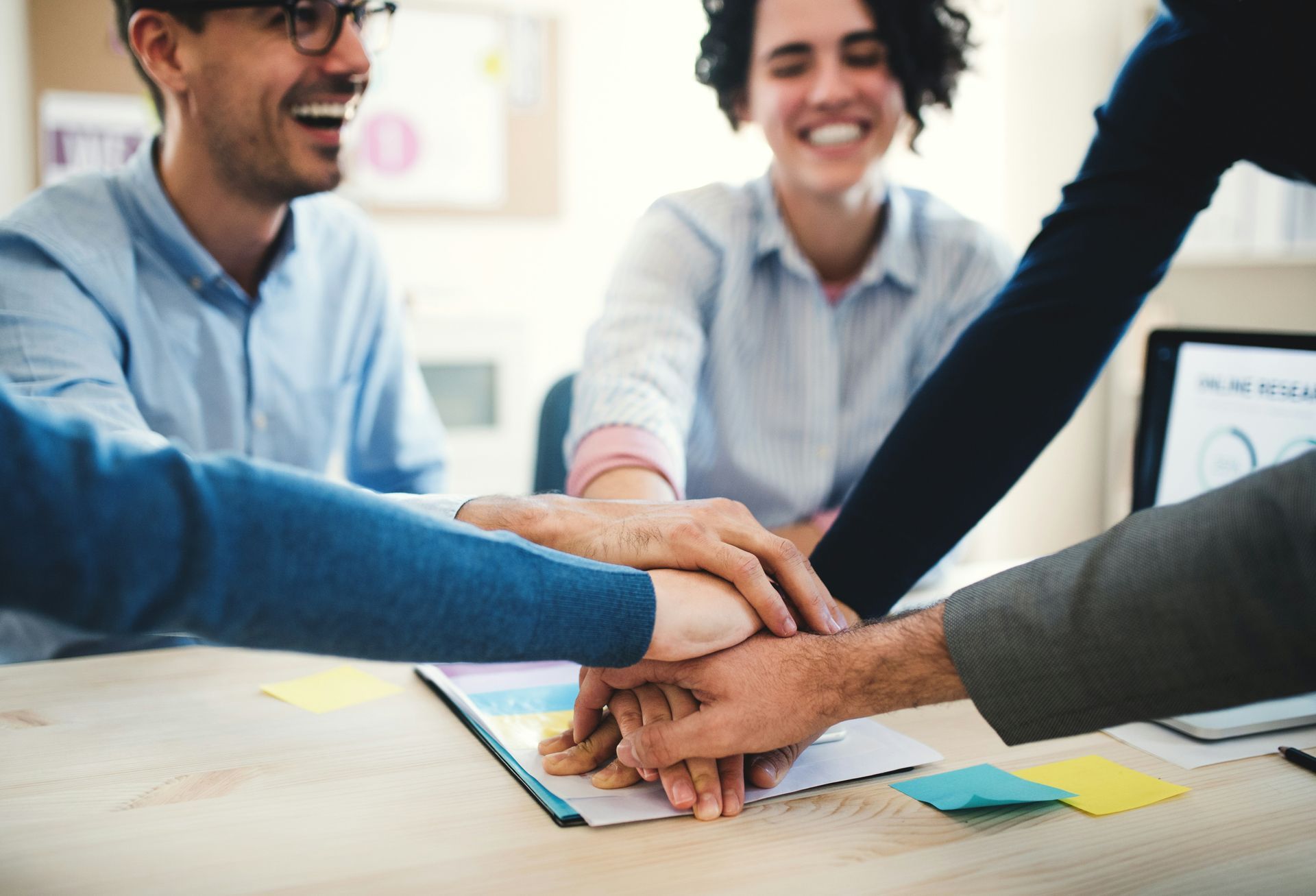Coworkers stacking hands in a team huddle at a desk with sticky notes and a laptop