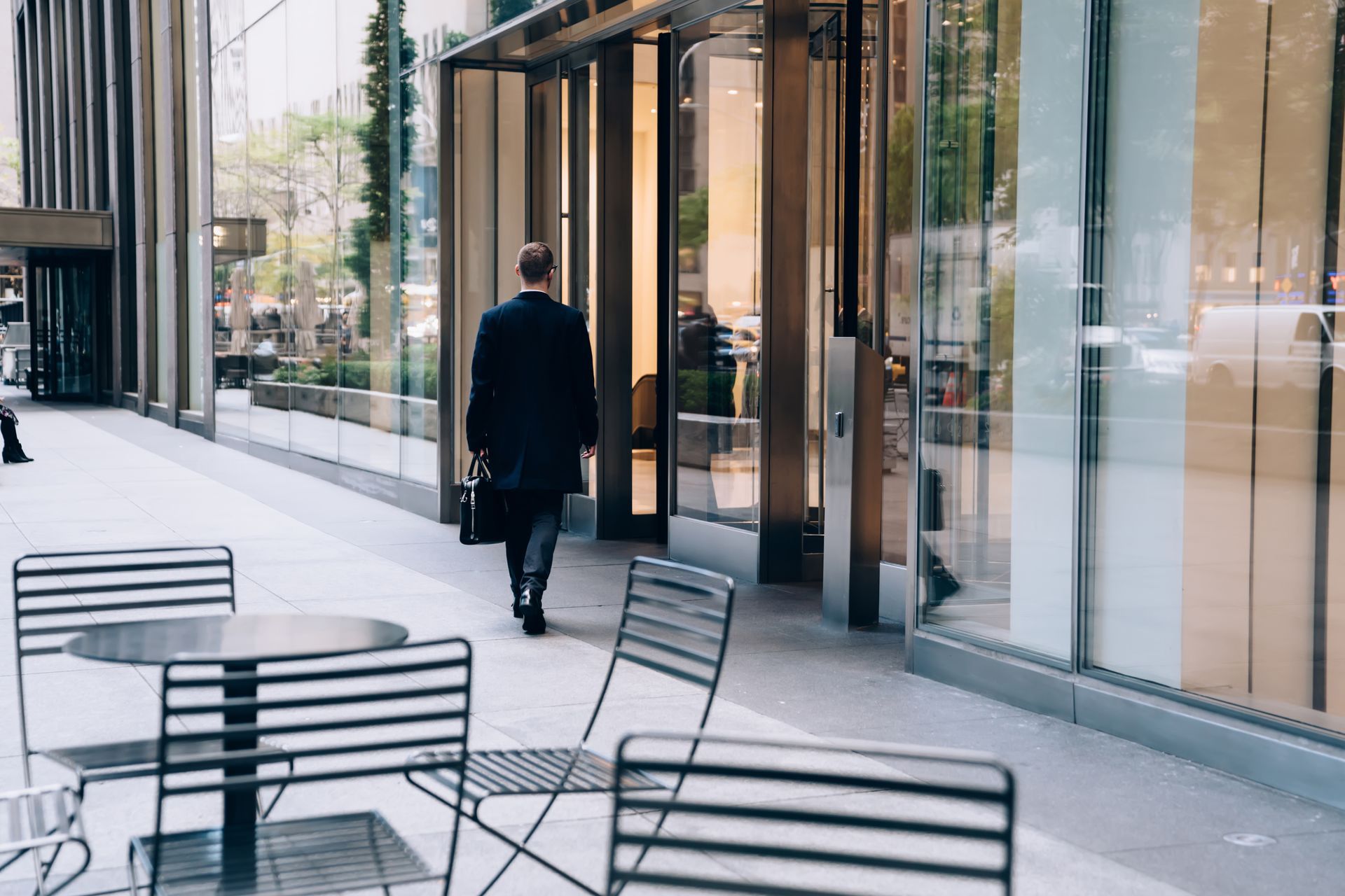 Person walking along a glass-fronted office terrace beside black metal café tables and chairs.