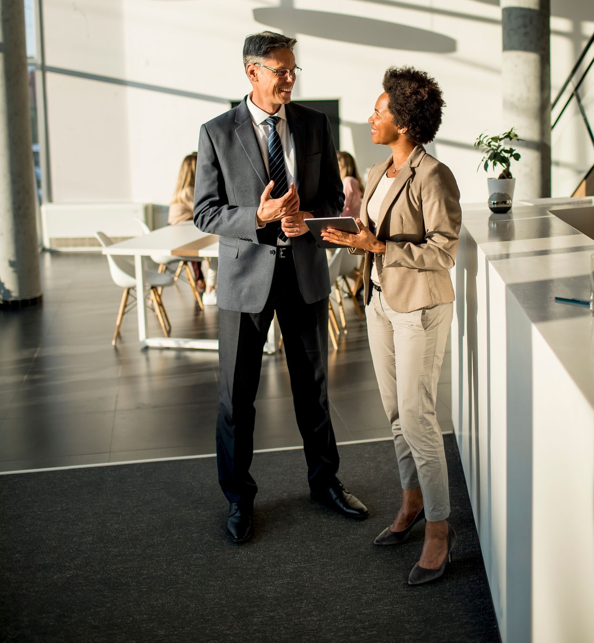 Two professionals talking in a bright modern office lobby, one in a dark suit and one in beige attire.