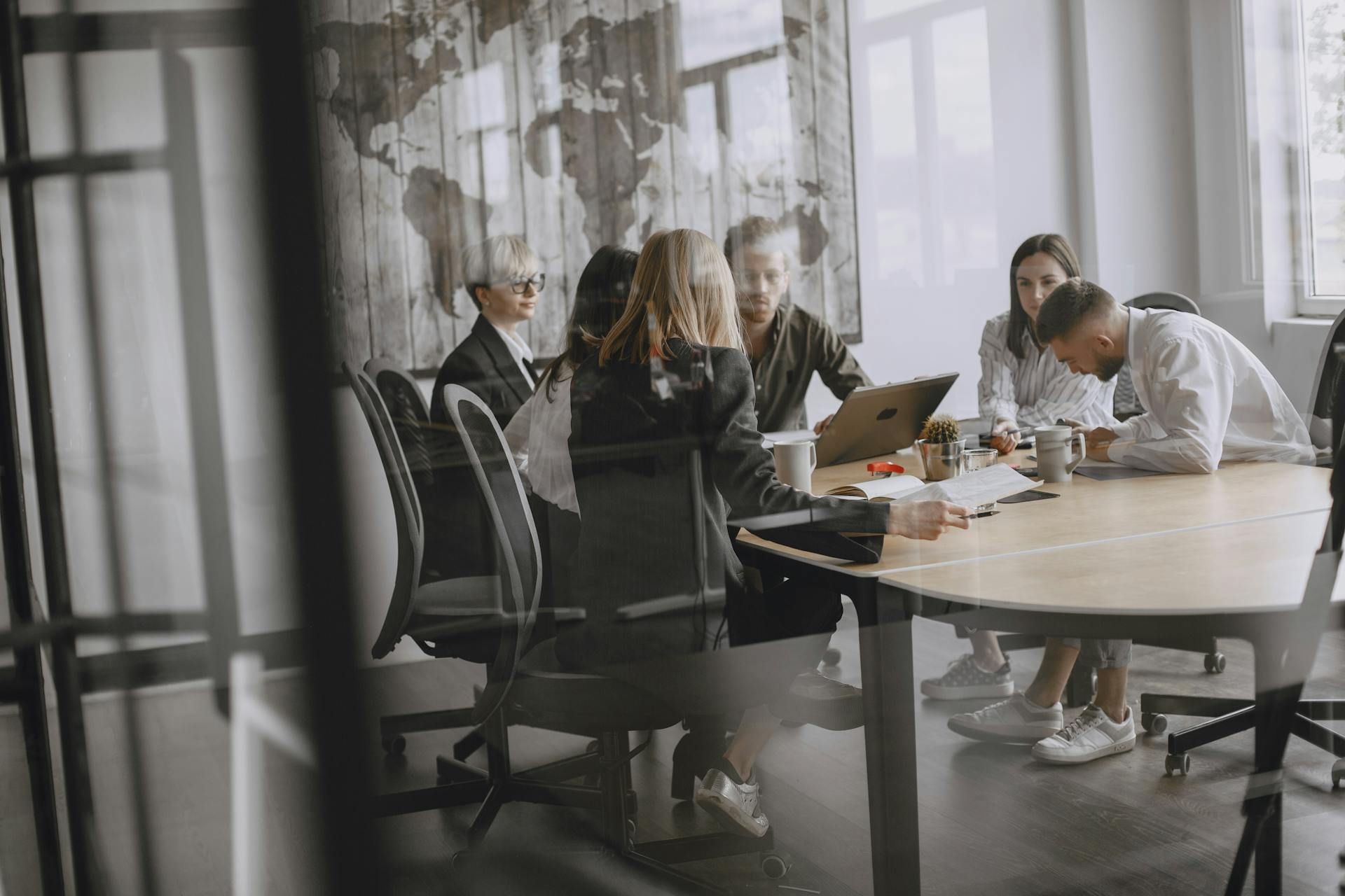 People in a modern meeting room gathered around a table, discussing a laptop and papers.