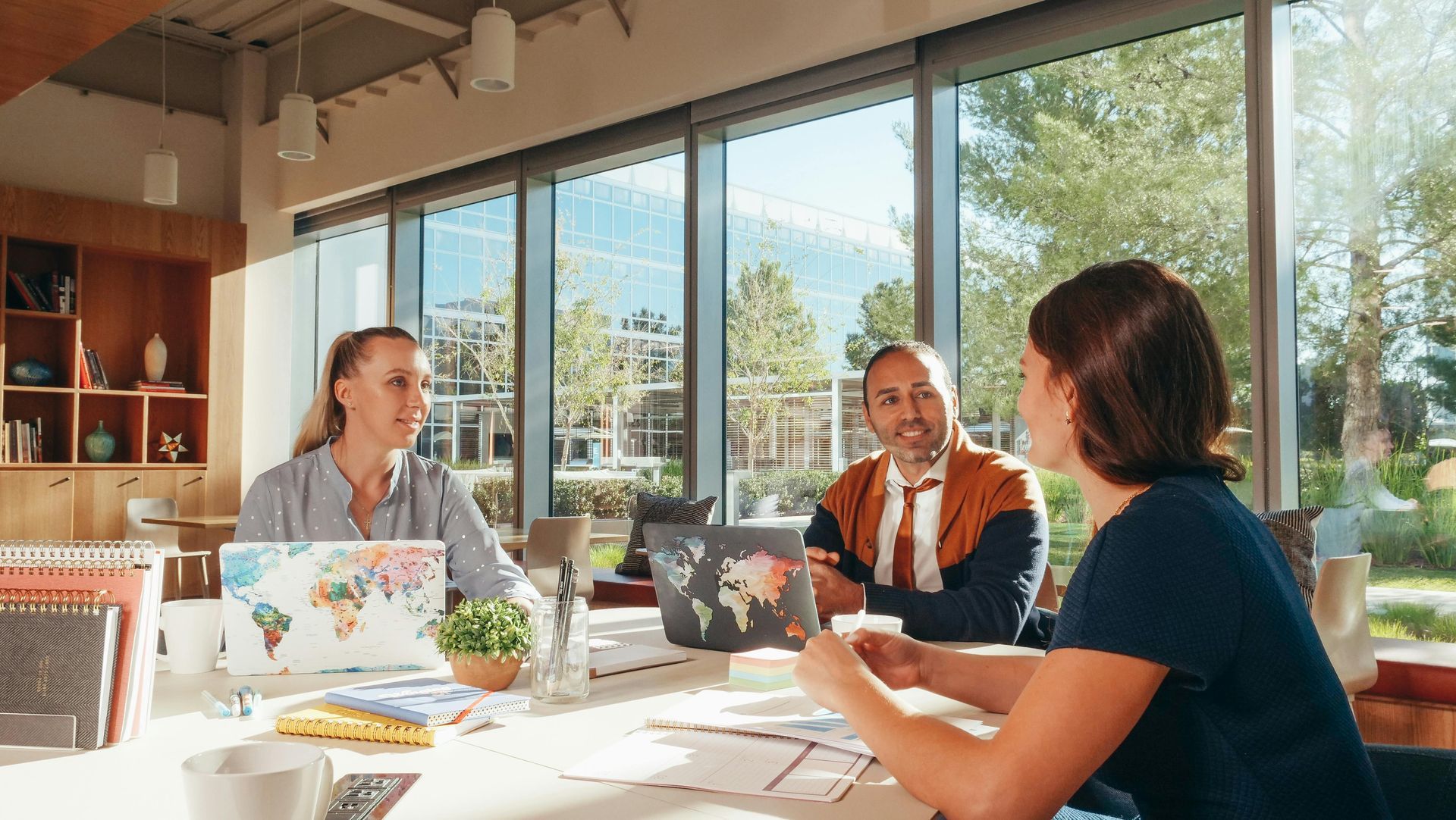Three people collaborating at a sunlit table in a modern office with laptops and papers.