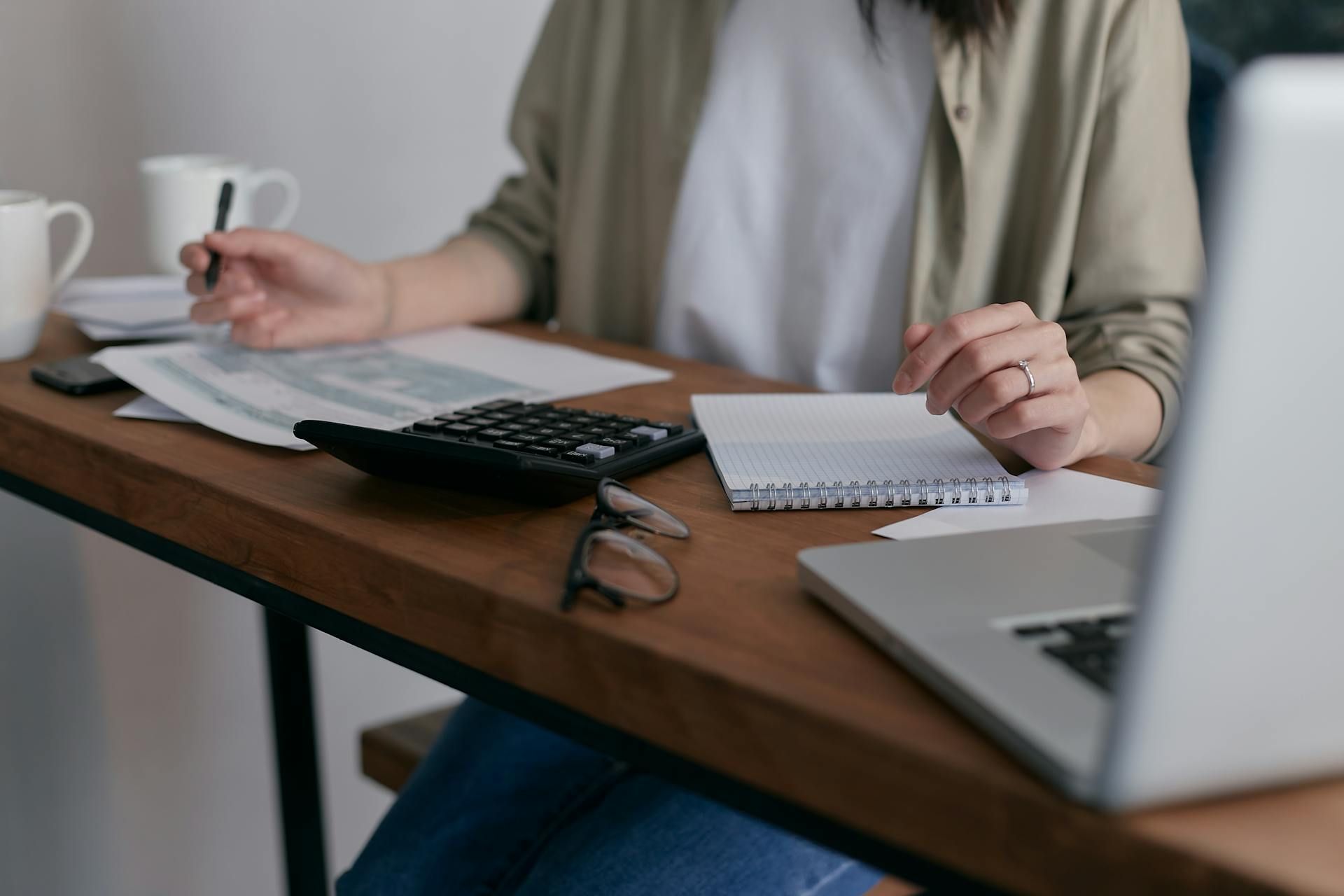 Person working at a desk with a calculator, notebook, glasses, laptop, and coffee mug
