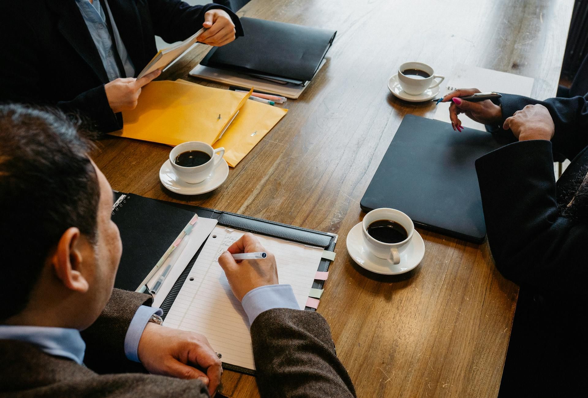 Team meeting at a wooden table with notebooks, coffee cups, and people taking notes