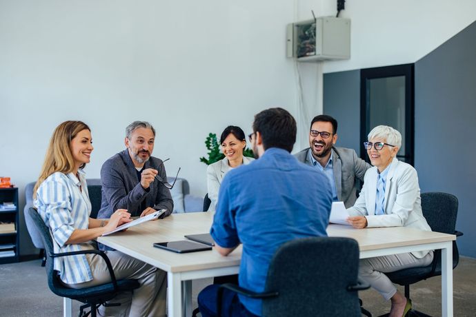 Group of coworkers in a meeting room, seated around a table and talking with laptops and notes.