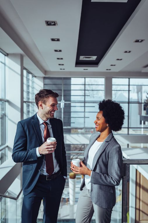 Two coworkers in business attire talking with coffee in a bright modern office lobby