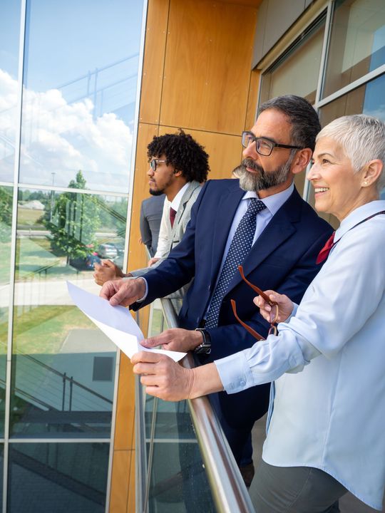 Three people reviewing documents on a balcony overlooking glass office buildings