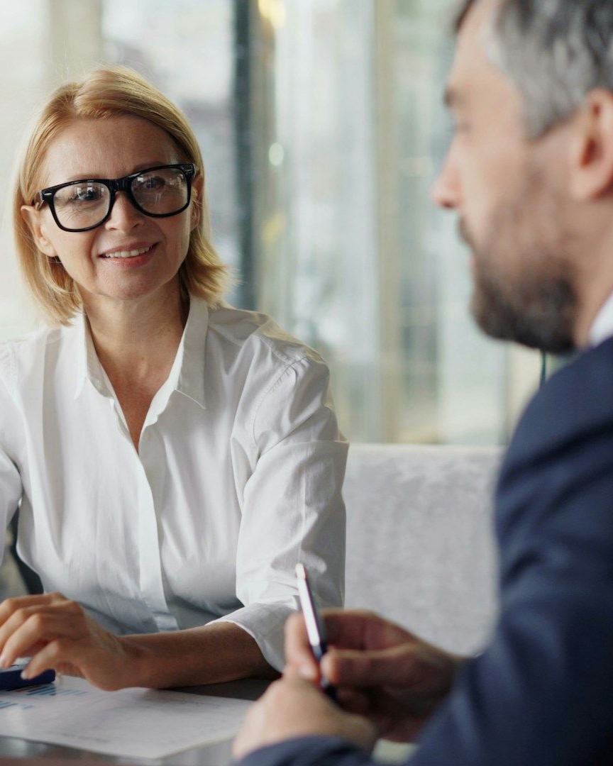 Two colleagues in a meeting, one smiling in glasses and white blouse, the other holding a pen.