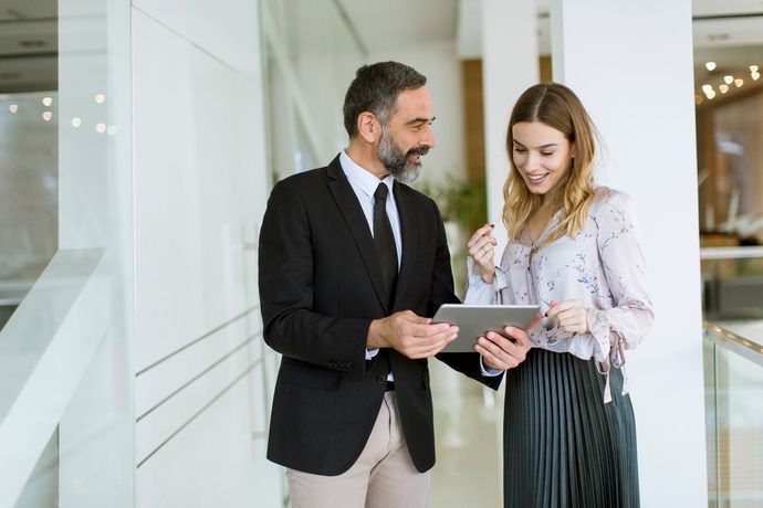 Two colleagues in business attire reviewing a tablet in a bright office hallway