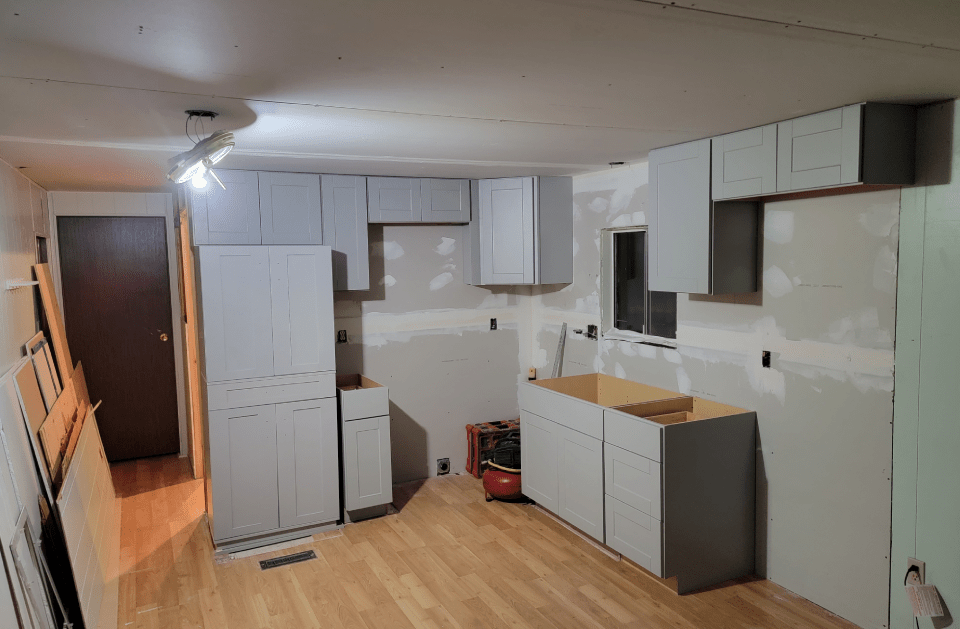 Kitchen renovation in progress: grey cabinets installed on drywall walls, hardwood floor.