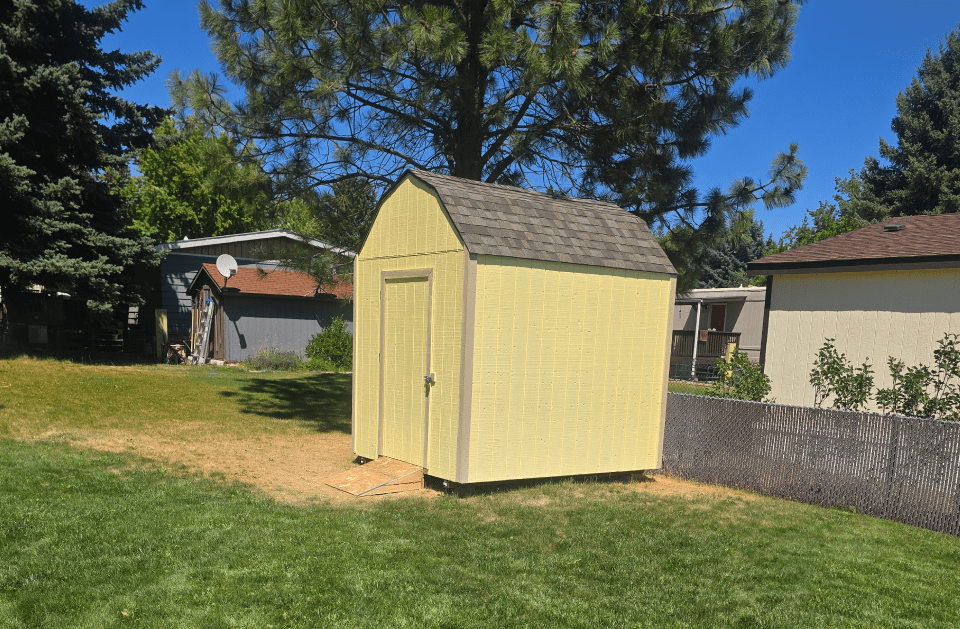 Yellow storage shed in a grassy yard, with a brown roof and a blue sky in the background.