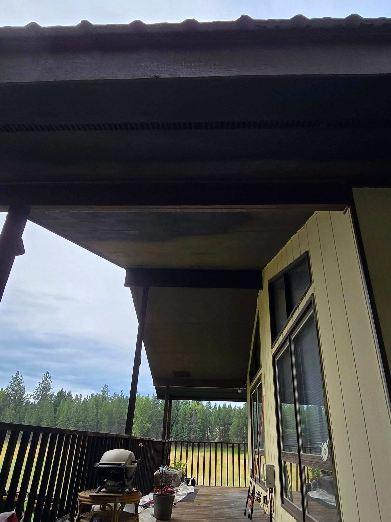 Covered porch with dark beams, a grill, and a forest in the background.