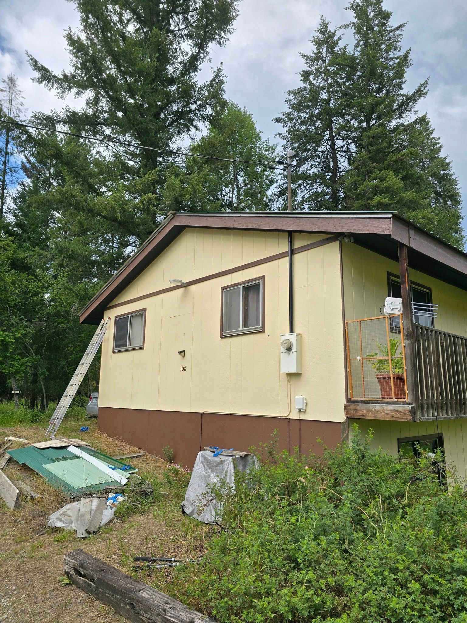 Tan house with brown trim and small balcony. Ladder leans against the side. Overgrown bushes in front.