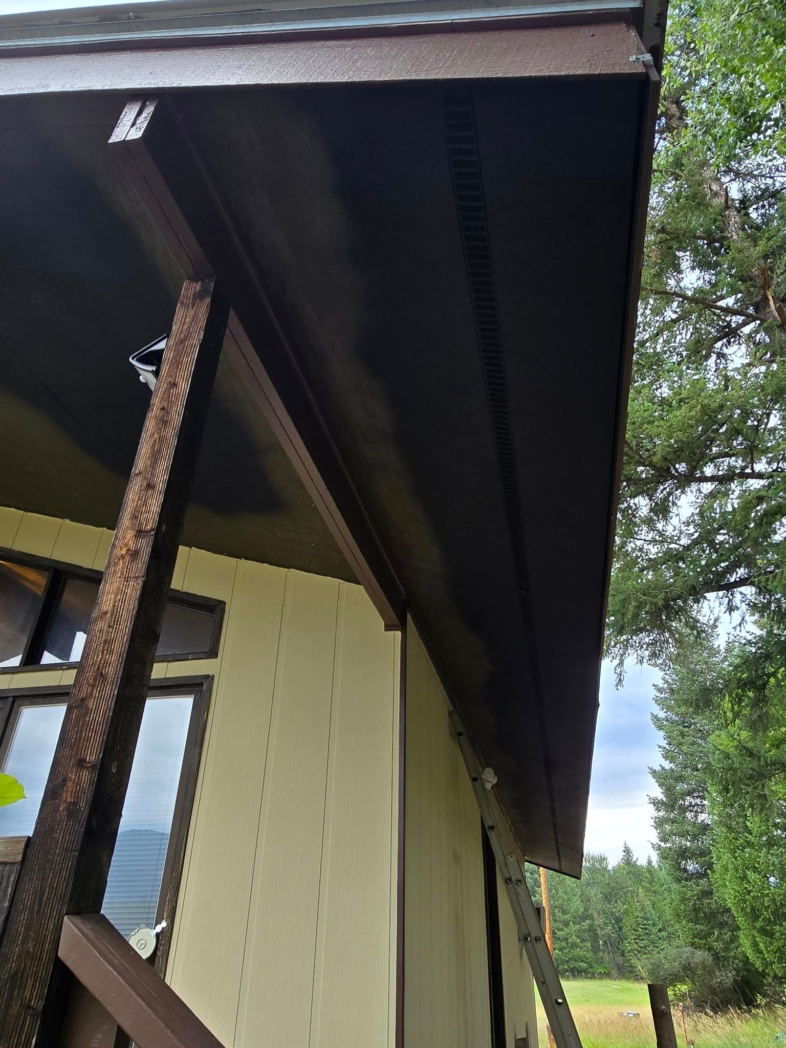 Brown wooden porch roof, supports, and siding on a cream-colored building. Trees and sky in background.