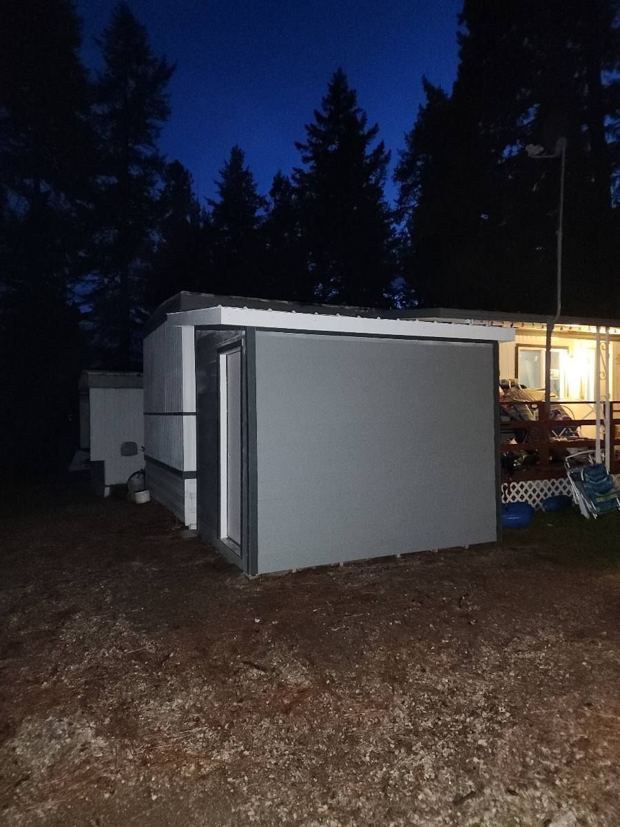 Gray shed with an open door at dusk, beside a house with a lit window, in a wooded area.