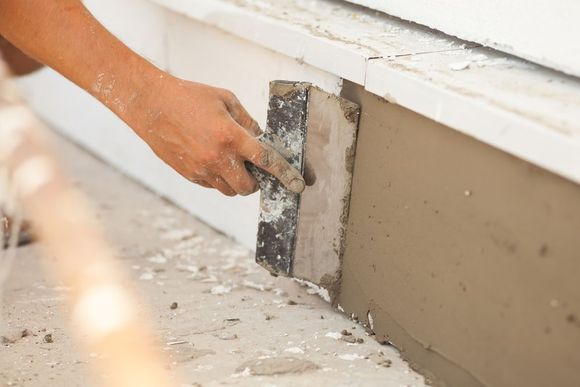 Person using a trowel to apply mortar on a concrete surface, near a white window frame.