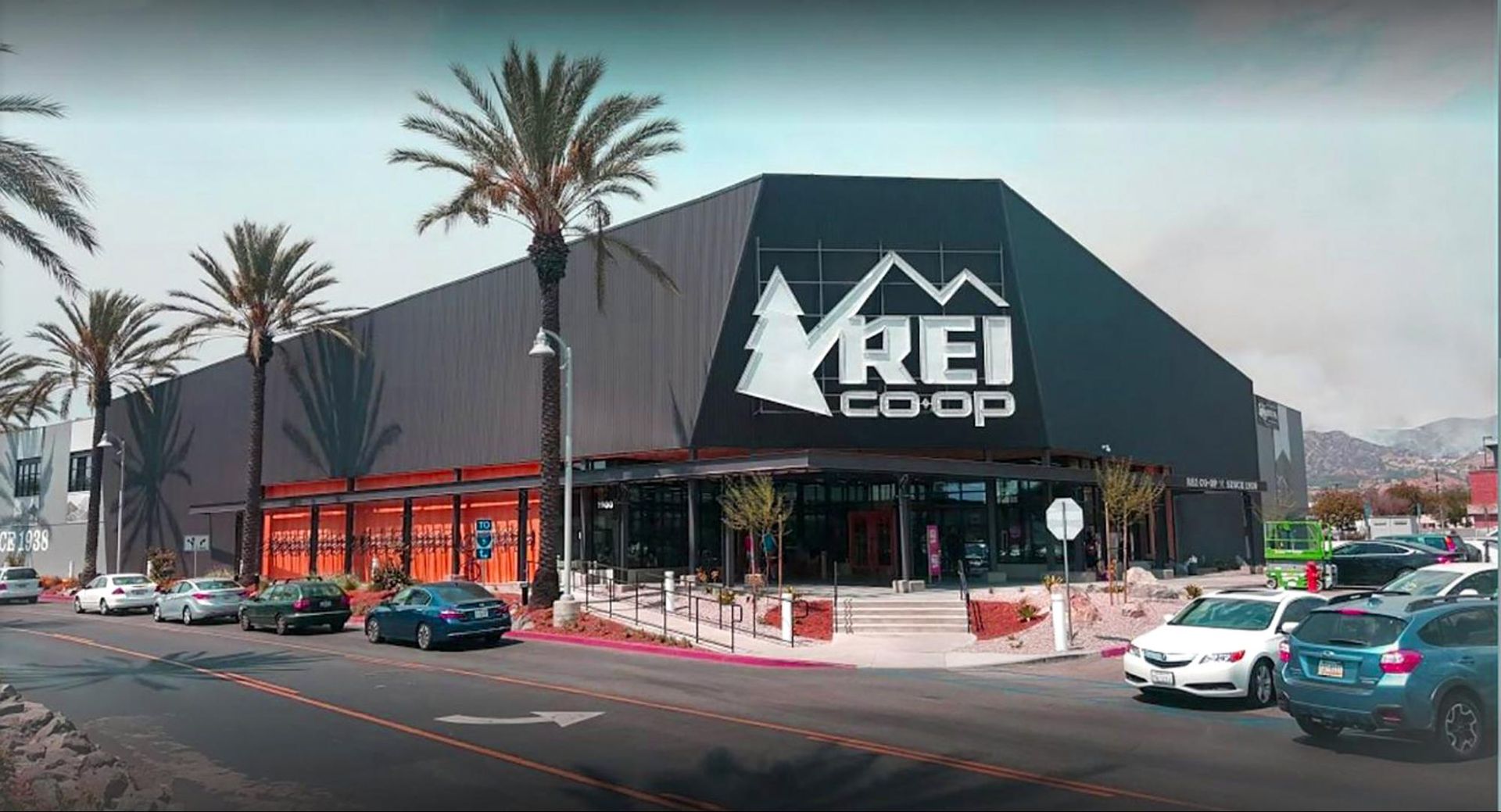 REI Co-op storefront with palm trees, parked cars, and mountains in the background