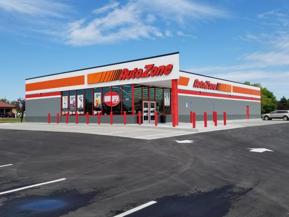 AutoZone store exterior with red-orange signage, glass entrance, and empty parking lot on a sunny day