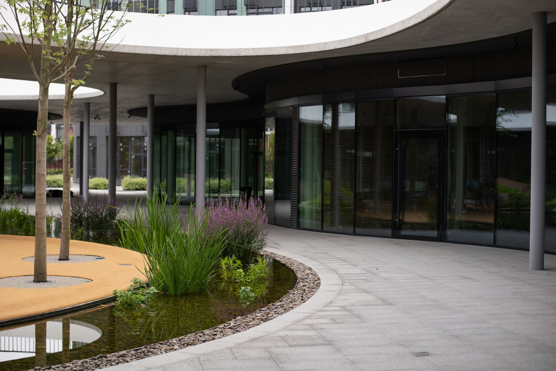 Modern glass office building entrance with curved walkway, landscaped plants, and outdoor seating area