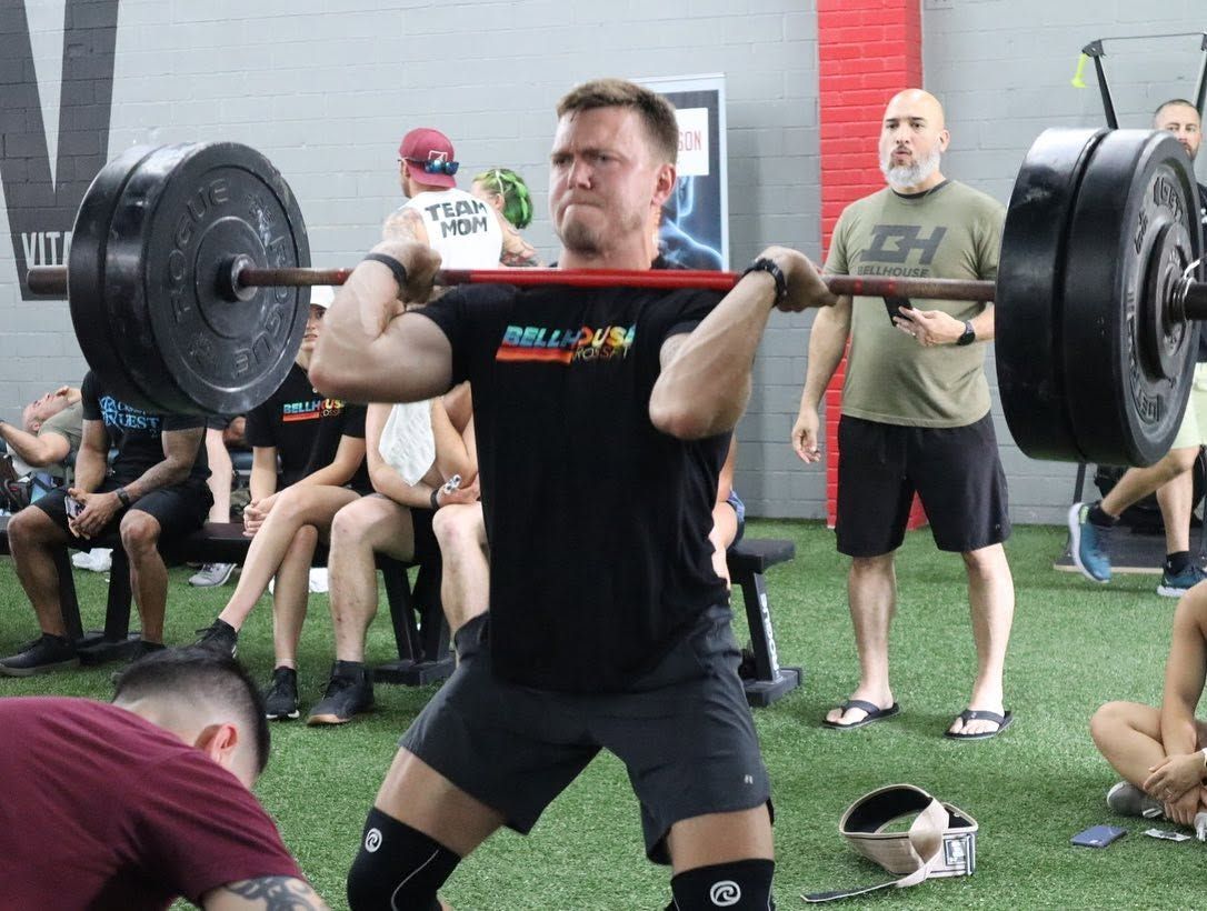 Man lifting a barbell overhead in a gym in Baytown, TX, with spectators seated behind him.