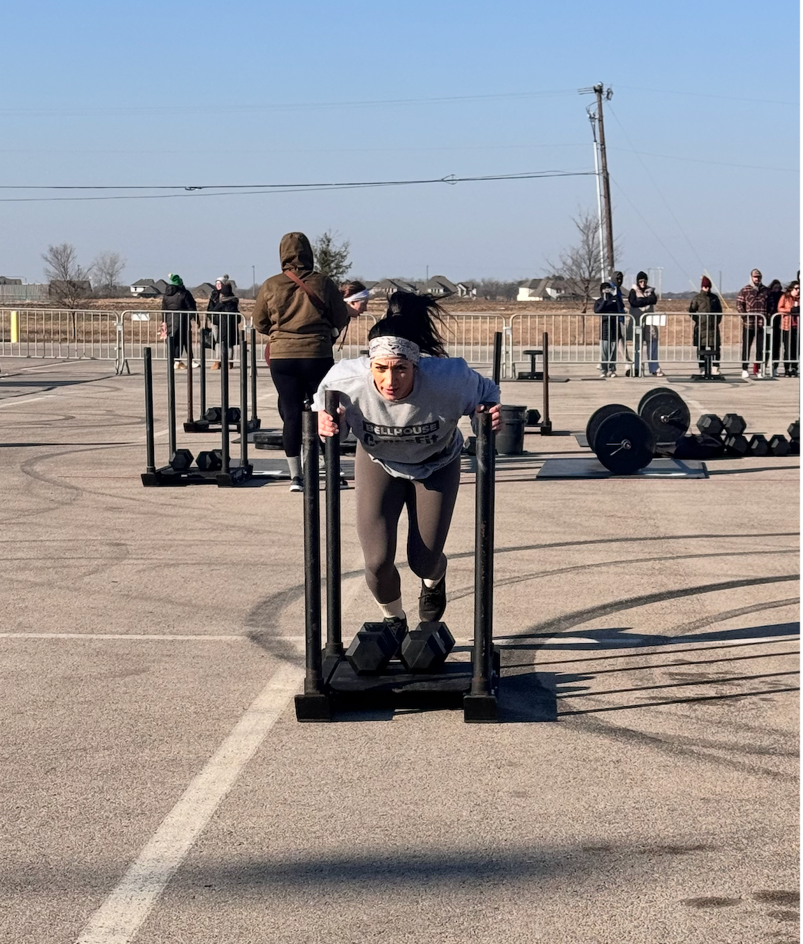 Person pushing a weighted sled on an outdoor lot, with gym in Baytown, TX equipment and people in the background.