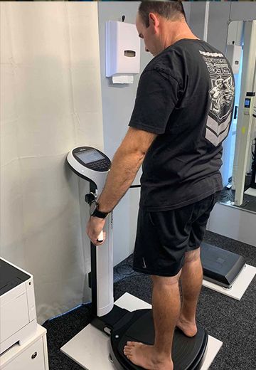 Person standing on a body composition scale in a gym, holding the handles, facing a wall-mounted display.