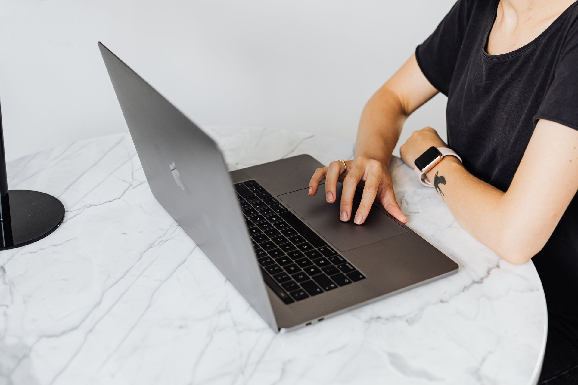 Person typing on a laptop at a white marble table, wearing a black shirt and smartwatch.