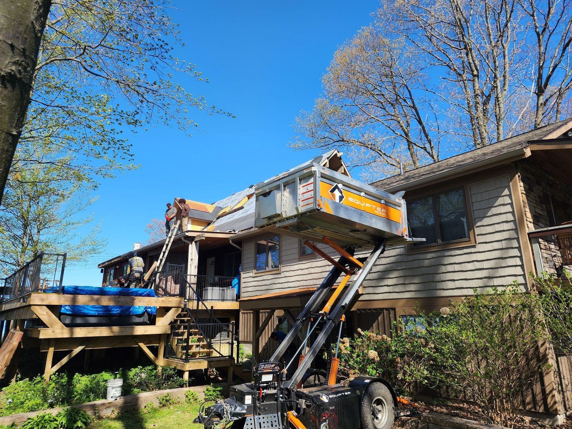 Dumpster lift loading debris onto a house roof for roof repair, with workers and bare trees around the home